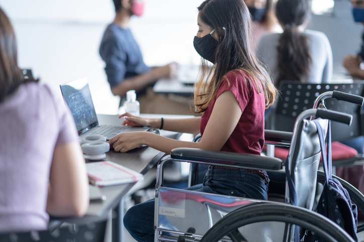 A female student of Asian ethnicity is seated on her wheelchair in her classroom. She is wearing a face mask to help prevent the spread of g