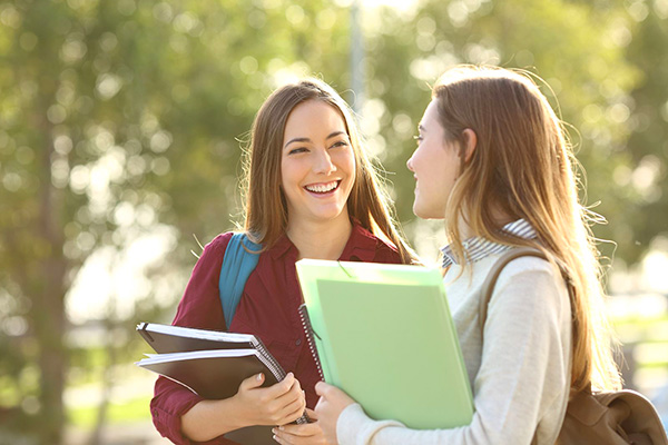 Two college-aged girls smiling toward each other, holding notebooks