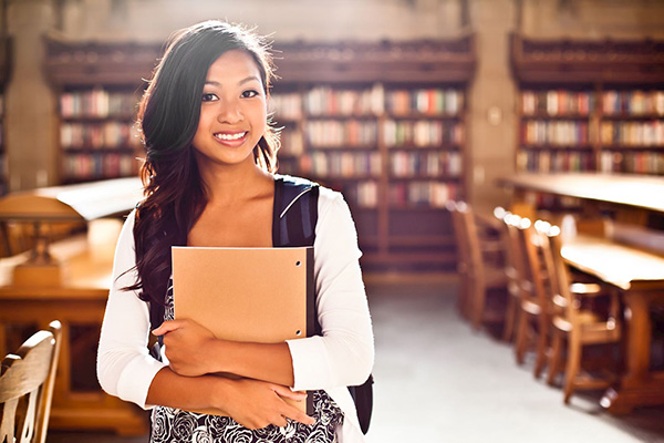 Attractive college-aged Asian girl holding notebook, standing in library