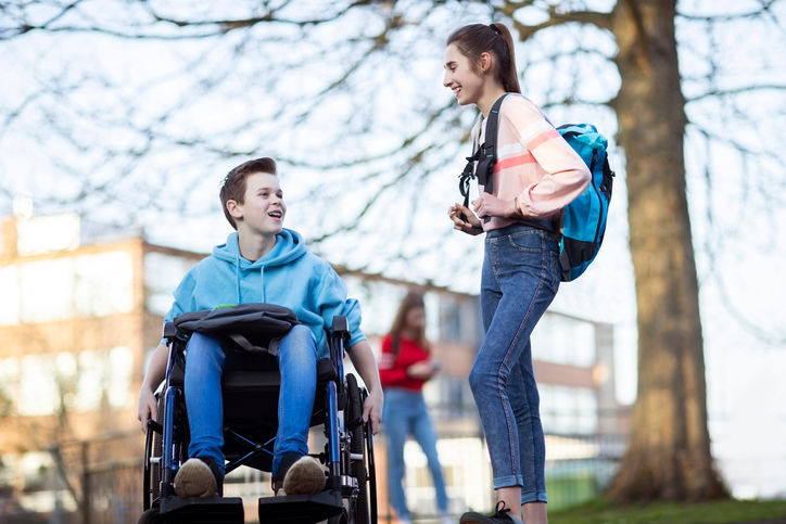 Teenage Boy In Wheelchair Talking With Female Friend As They Leave High School                                                              