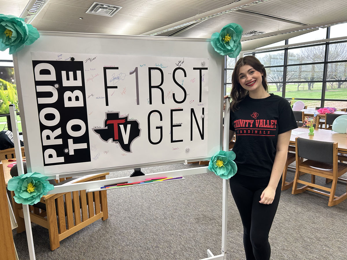 a first generation student standing next to a white board with the tvcc first gen logo and text "proud to be a first gen"