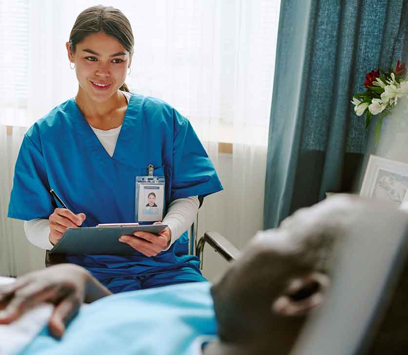 Female caretaker sitting next to man in hospital bed and taking notes on clipboard