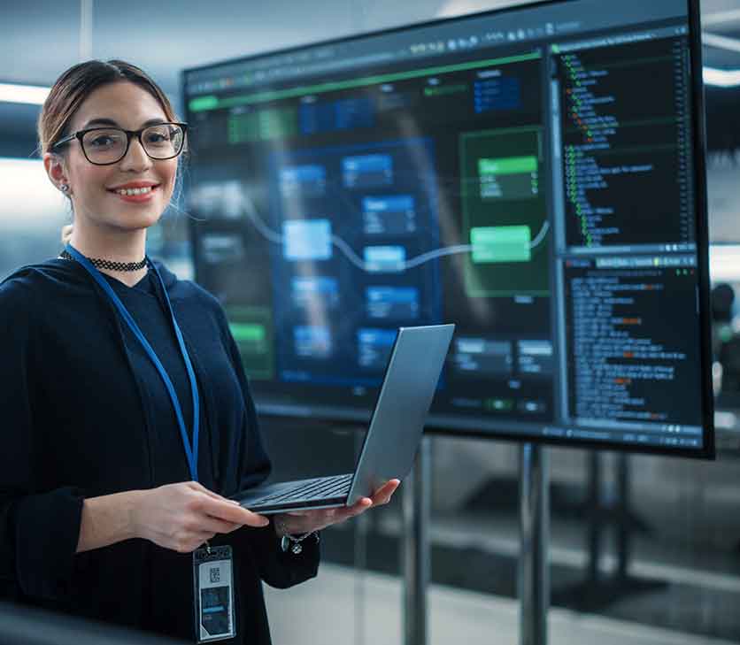 Woman smiling and holding laptop in front of a TV monitor showing programming