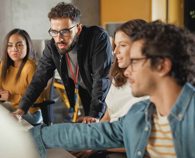 Male instructor guiding a group of students huddled around him on a computer